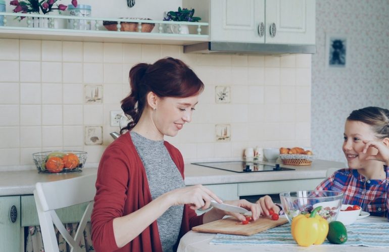 Cheerful happy mother cutting tomatoes while cooking and talking to her smiling daughter feeding mom in the kitchen at home. Family, cook, and people concept