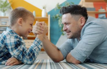 caring father and active son enjoying arm wrestling on floor at home laughing having fun together. People, sports activities and house concept.